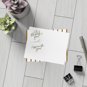 May include: A white rectangular card with gold vertical stripes and the dark green cursive text We thank you! and Appreciate you! The card is positioned near two black metal binder clips and a grey pen, with two potted succulents nearby.