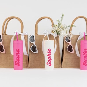 Puede incluir: Tres bolsas de yute con cinta blanca y gafas de sol. Cada bolsa tiene un vaso personalizado con un nombre en rosa. Un vaso es blanco. Flores en una bolsa.