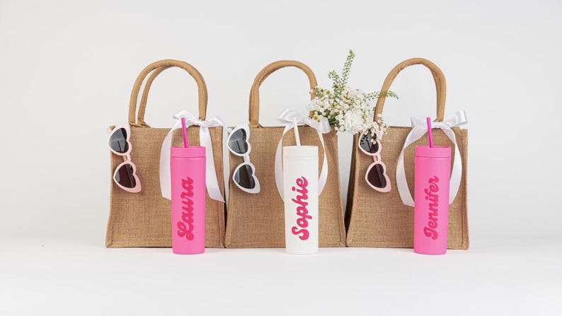 Puede incluir: Tres bolsas de yute con cinta blanca y gafas de sol. Cada bolsa tiene un vaso personalizado con un nombre en rosa. Un vaso es blanco. Flores en una bolsa.