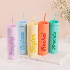 Puede incluir: Cinco vasos de colores con pajitas, cada uno en un tono pastel diferente: lavanda, azul claro, amarillo, naranja y turquesa. Cada vaso tiene un nombre impreso en una fuente retro a juego: Hayley, Chelsea, Taylor, Rachel y Amanda.