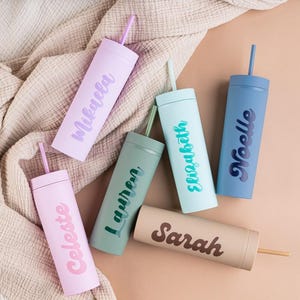 Puede incluir: Una variedad de vasos personalizados en colores pastel, cada uno con una pajita a juego. Los vasos son de color rosa claro, lavanda, verde salvia, azul claro, beige y azul marino, cada uno con un nombre en un color complementario.