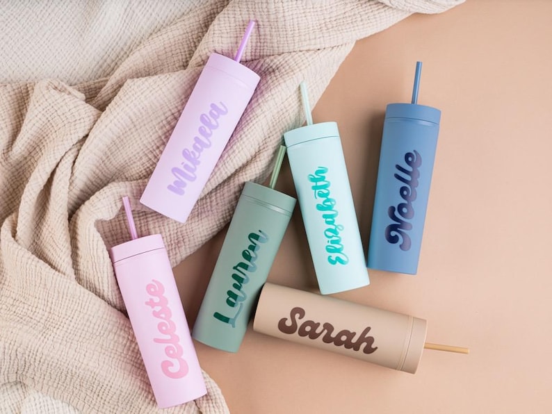 Puede incluir: Una variedad de vasos personalizados en colores pastel, cada uno con una pajita a juego. Los vasos son de color rosa claro, lavanda, verde salvia, azul claro, beige y azul marino, cada uno con un nombre en un color complementario.