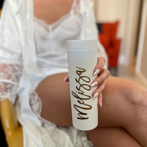 Puede incluir: Un vaso blanco con el nombre "Melissa" en escritura dorada es sostenido. El vaso tiene una pajita blanca. La persona lleva una bata blanca con ribete de encaje.