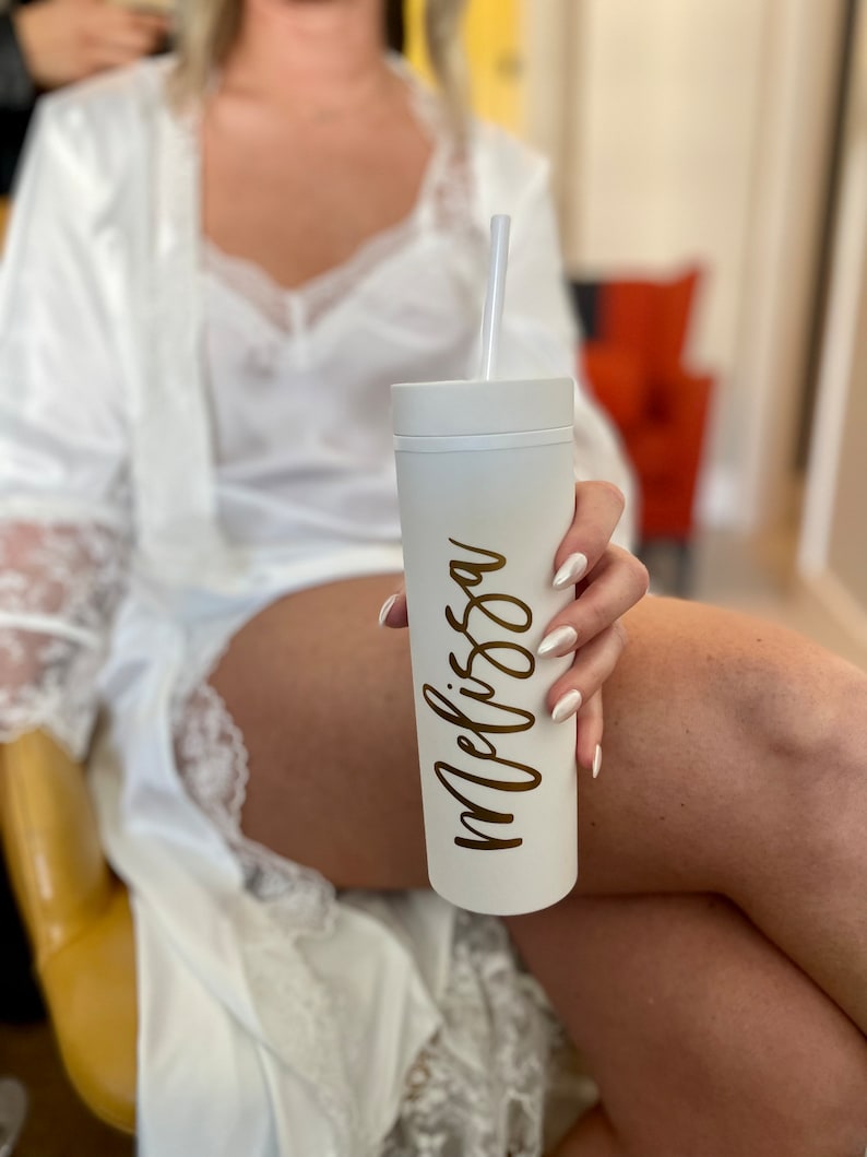 Puede incluir: Un vaso blanco con el nombre "Melissa" en escritura dorada es sostenido. El vaso tiene una pajita blanca. La persona lleva una bata blanca con ribete de encaje.