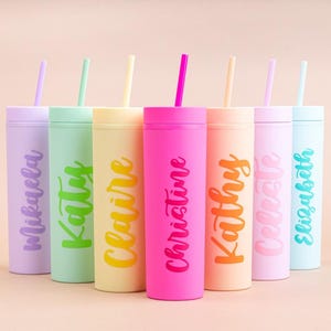 Puede incluir: Una colecci&oacute;n de siete vasos de colores con tapas y pajitas. Cada vaso es de un color pastel diferente y tiene un nombre escrito en una fuente de escritura a juego. Los vasos est&aacute;n dispuestos en fila sobre un fondo rosa claro.