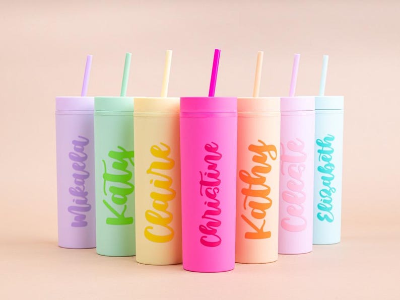 Puede incluir: Una colecci&oacute;n de siete vasos de colores con tapas y pajitas. Cada vaso es de un color pastel diferente y tiene un nombre escrito en una fuente de escritura a juego. Los vasos est&aacute;n dispuestos en fila sobre un fondo rosa claro.