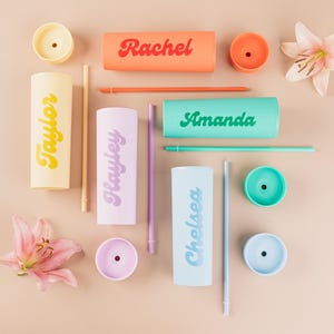 Puede incluir: Una vista cenital de vasos personalizados en varios colores, cada uno con un nombre impreso en el lateral. Los nombres incluyen "Rachel", "Amanda", "Taylor", "Hayley" y "Chelsea". La imagen tambi&eacute;n incluye tapas, pajitas y flores a juego.