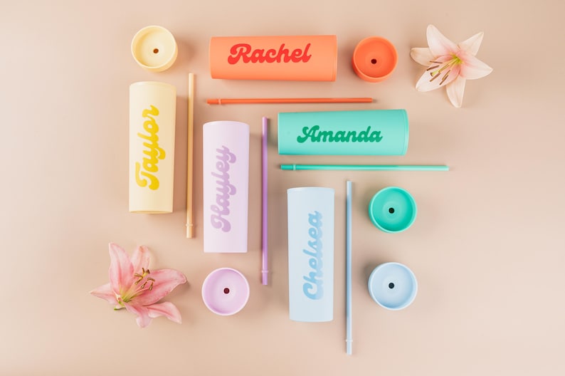 Puede incluir: Una vista cenital de vasos personalizados en varios colores, cada uno con un nombre impreso en el lateral. Los nombres incluyen "Rachel", "Amanda", "Taylor", "Hayley" y "Chelsea". La imagen tambi&eacute;n incluye tapas, pajitas y flores a juego.