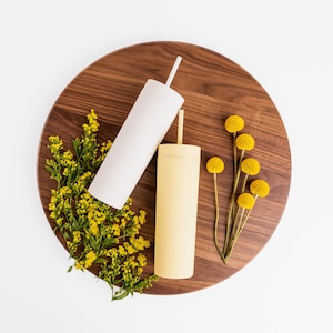 Puede incluir: Dos vasos aislados, uno blanco y otro amarillo p&aacute;lido, con pajitas, descansan sobre una tabla de madera oscura. Flores amarillas y vegetaci&oacute;n se disponen alrededor de los vasos. Los vasos tienen un dise&ntilde;o moderno y minimalista.