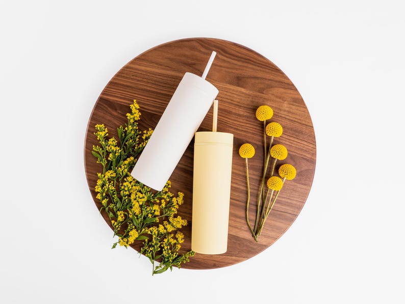 Puede incluir: Dos vasos aislados, uno blanco y otro amarillo p&aacute;lido, con pajitas, descansan sobre una tabla de madera oscura. Flores amarillas y vegetaci&oacute;n se disponen alrededor de los vasos. Los vasos tienen un dise&ntilde;o moderno y minimalista.