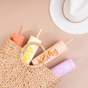 Puede incluir: Un bolso tote de paja tejida contiene cuatro vasos personalizados en tonos melocot&oacute;n, amarillo, coral y lavanda. Cada vaso tiene una pajita a juego y un nombre en fuente cursiva. Un sombrero para el sol blanco descansa cerca.
