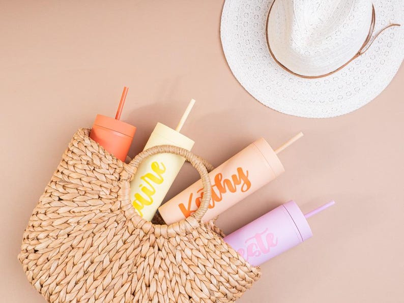 Puede incluir: Un bolso tote de paja tejida contiene cuatro vasos personalizados en tonos melocot&oacute;n, amarillo, coral y lavanda. Cada vaso tiene una pajita a juego y un nombre en fuente cursiva. Un sombrero para el sol blanco descansa cerca.