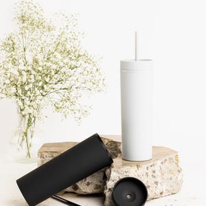 Puede incluir: Un vaso blanco con pajita, un vaso negro y una tapa negra se exhiben sobre una superficie de piedra. Un jarr&oacute;n con flores blancas est&aacute; en el fondo. Los vasos son cil&iacute;ndricos y parecen ser de metal.