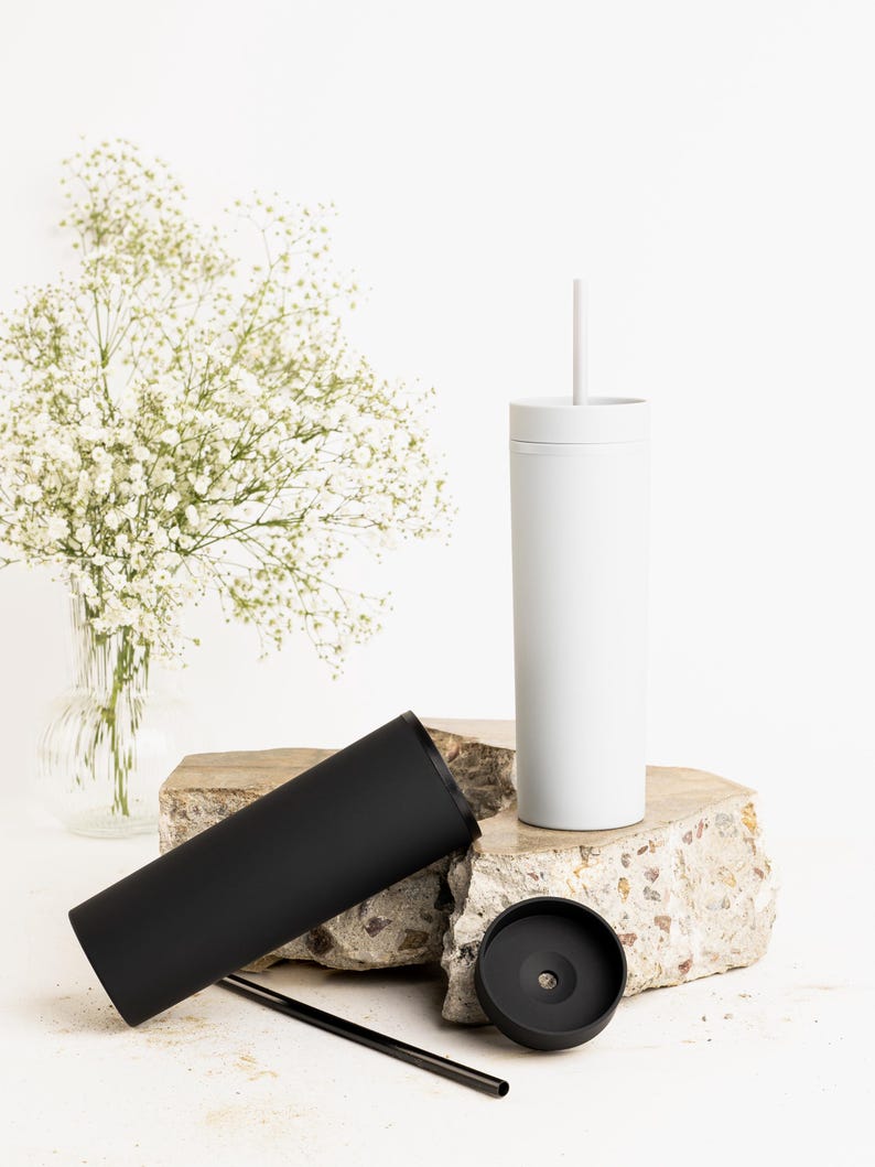 Puede incluir: Un vaso blanco con pajita, un vaso negro y una tapa negra se exhiben sobre una superficie de piedra. Un jarr&oacute;n con flores blancas est&aacute; en el fondo. Los vasos son cil&iacute;ndricos y parecen ser de metal.