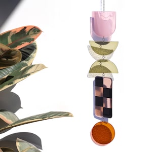 Puede incluir: Un móvil colgante vertical compuesto por cinco formas geométricas distintas unidas por anillos de metal. Las piezas incluyen un arco rosa translúcido, dos semicírculos dorados, un panel rectangular con un patrón de tablero de ajedrez negro y cobre, y un disco circular naranja en la base.