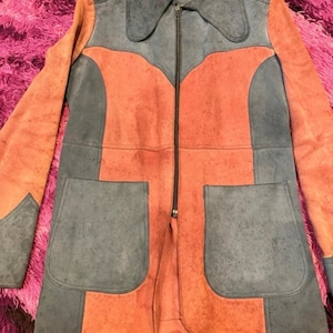 May include: A color-blocked jacket featuring a combination of slate blue and burnt orange panels. The jacket has a full-length front zipper, a pointed collar, and two large patch pockets on the front. The design includes long sleeves.