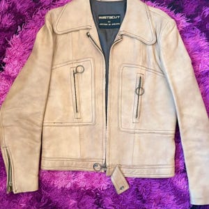 May include: A beige leather jacket featuring a wide collar, a central zip, and two vertical zipped pockets on the chest. The interior label reads Mastacut by Austin of England, Made in England. The sleeves have zipped cuffs and a belt tab.