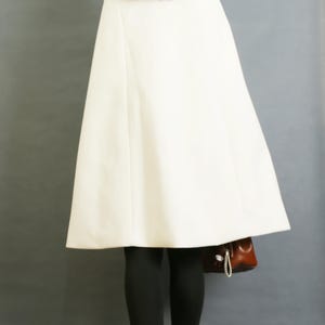 Pode incluir: Uma capa de cor branco-sujo lisa com uma textura suave, apresentando uma silhueta larga e evas&ecirc; que cai dos ombros. A pe&ccedil;a &eacute; usada sobre leggings pretas e combinada com uma mala castanha e botas vermelho-escuras, vista de costas.