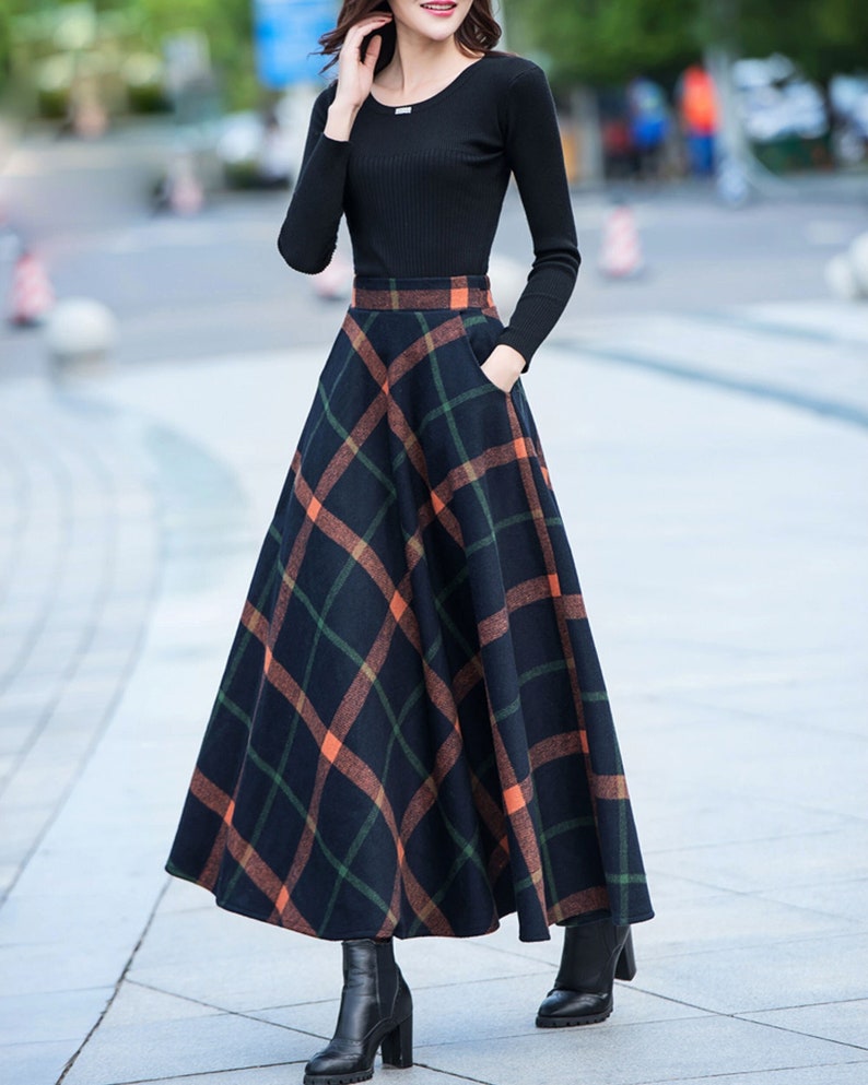 Peut inclure: Une chemise noire &agrave; manches longues avec une jupe &agrave; carreaux noirs et orange. La jupe est trap&egrave;ze et arrive aux chevilles. La femme porte des bottes &agrave; talons noires.