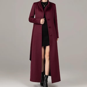 May include: A long, burgundy coat with a high collar and button closure. The coat has a tailored fit and a split front, reaching to the ankles. The model is wearing black boots. The coat is made of a soft, flowing fabric.