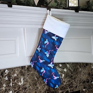Può includere: Un calzino di Natale blu e rosa con un polsino bianco. Il calzino presenta un motivo ripetuto di dragoni con ali rosa e blu.