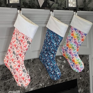 Può includere: Tre calze di Natale con polsini bianchi appese a un camino. Le calze sono decorate con diversi motivi: rosa e bianco, blu e bianco e arcobaleno.
