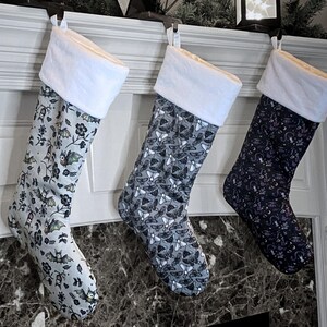 May include: Three Christmas stockings with different spooky designs. The stocking on the left has a white background with bats, flowers, and skulls. The middle stocking has a gray background with moths. The stocking on the right has a black background with a purple, green, and white design.