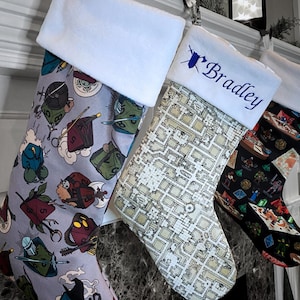 May include: Three Christmas stockings with white cuffs hang from a mantel. The stockings feature various patterns, including dice, maps, and fantasy-themed illustrations. One stocking is personalized with the name "Bradley" in blue embroidery.