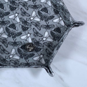 May include: Hexagonal gray fabric tray with a repeating pattern of black and white moths. The tray has raised edges and is designed to hold small items. A silver-colored metal object rests inside the tray.