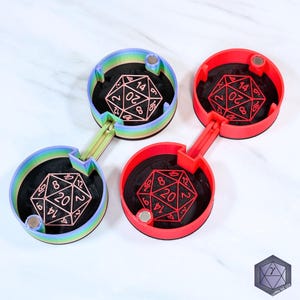 May include: Four dice holders in the image. Two are red, and two are rainbow-colored. Each holder has a black interior and a D20 die. The holders are connected by a hinge. The dice are red and copper-colored.