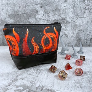 May include: A black and red dice bag with an octopus design. The bag is made of fabric and has a leather bottom. There are several dice on a gray surface next to the bag.