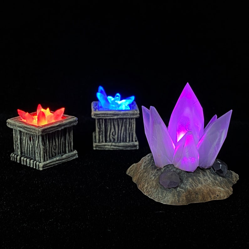 May include: Three glowing crystal formations, each emitting a different color of light. The crystals are displayed on a dark surface. The first crystal is red and is in a small, square, gray container. The second crystal is blue and is in a small, square, gray container. The third crystal is purple and is on a gray, rock-like base.
