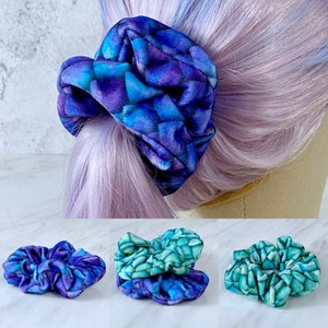 May include: Three colorful hair scrunchies with a mermaid scale pattern. The scrunchies are in shades of purple, blue, and green.