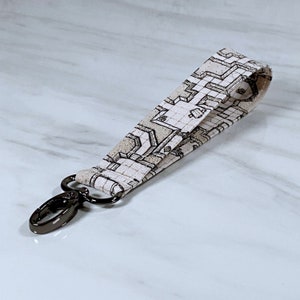 Op de afbeelding: Een stoffen polsband met een geometrisch patroon in beige en grijstinten. De lanyard heeft een zwarte metalen draaibare sluiting voor het bevestigen van sleutels of andere kleine items. De stof is gevouwen en gestikt.