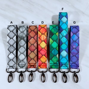 May include: Seven colorful keychains with a dragon scale pattern. The keychains are made of fabric and have a silver metal clasp. The colors of the keychains are gray, brown, orange, green, blue, and purple.