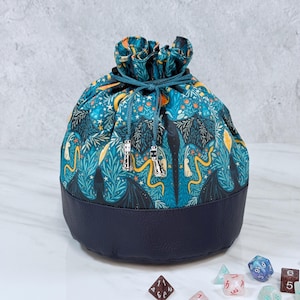 May include: A round, drawstring dice bag with a dark blue leather bottom and a patterned top. The top features a teal background with a repeating design of dragons, stars, and foliage. Silver tassels hang from the drawstring closure. Dice are scattered around the bag.