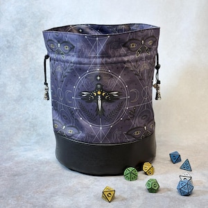 May include: A purple dice bag with a black leather base. The bag features a celestial design with a central raven illustration, geometric shapes, and eye motifs. Black drawstrings with silver tassels are visible.