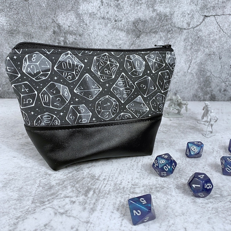 May include: A black zippered pouch with a white and gray dice pattern. The pouch has a black leather bottom. There are several blue dice scattered around the pouch.