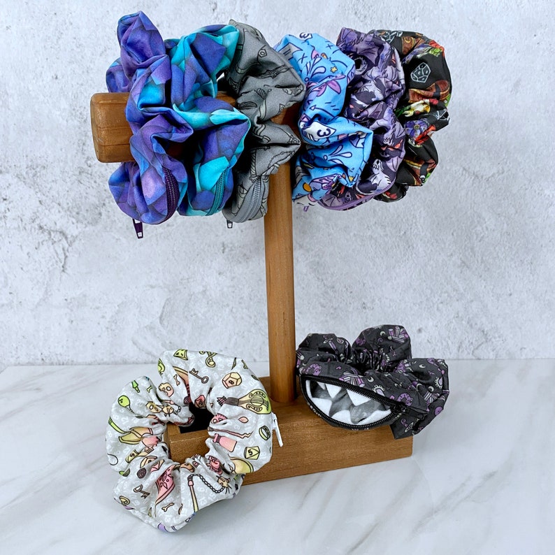 May include: A set of five colorful scrunchies displayed on a wooden stand. The scrunchies are made of fabric and have various patterns, including a purple and blue floral design, a black and white skull design, and a blue and white geometric design.