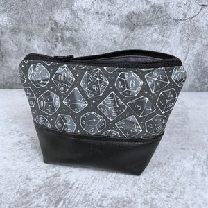 May include: Black zippered pouch with a white dice pattern on a black background. The pouch has a black leather bottom.
