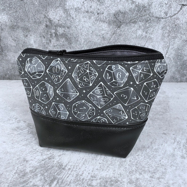 May include: Black zippered pouch with a white dice pattern on a black background. The pouch has a black leather bottom.