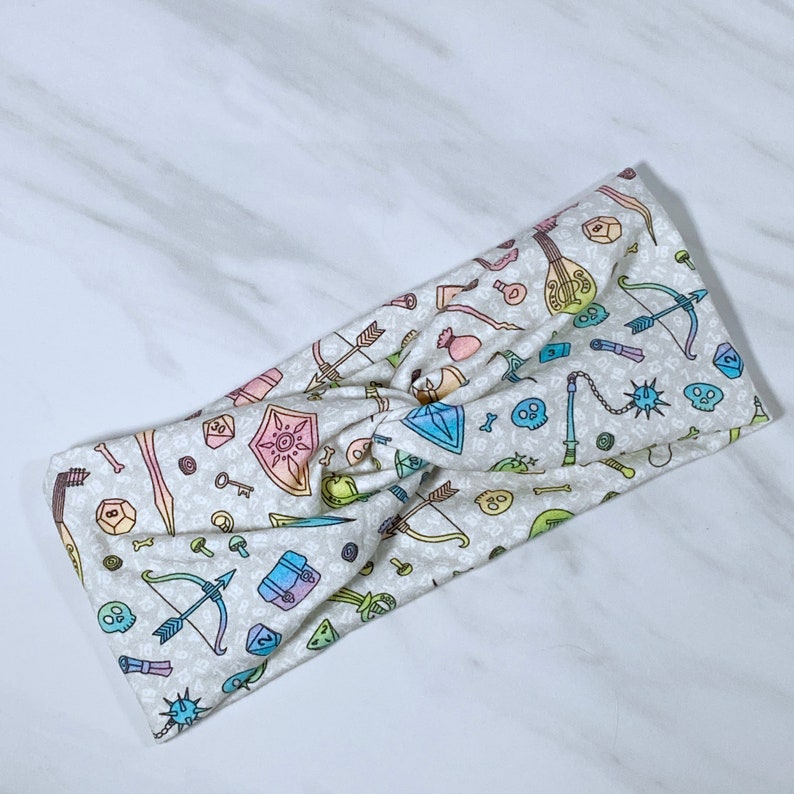 Puede incluir: Una diadema blanca con un estampado colorido de varios art&iacute;culos de tem&aacute;tica fant&aacute;stica y de juegos, incluyendo dados, espadas y un la&uacute;d.
