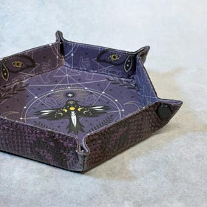 May include: A square, purple and black leather dice tray with a decorative interior. The interior features a celestial design with a central bird-like figure, moons, and geometric shapes. The exterior has a snakeskin pattern.