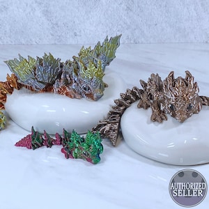 Könnte beinhalten: Drei kleine, farbige, 3D-gedruckte Drachenfiguren. Die Drachen sind auf weißen Keramiksockeln platziert. Die Drachen sind grün, rot und braun mit schillernden Schuppen.