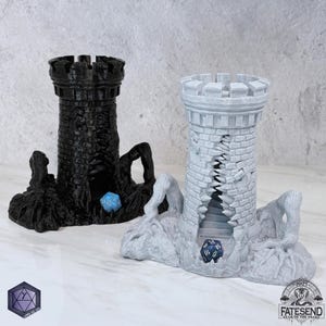 May include: Two dice towers, one black and one gray, resembling castle turrets. Each tower has a dice-rolling chamber and is set on a textured base. A blue die is visible in each tower. The towers are designed for tabletop gaming.