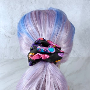 May include: A black hair scrunchie with a colorful print of cartoon monsters. The scrunchie is tied around a ponytail of purple and blue hair.