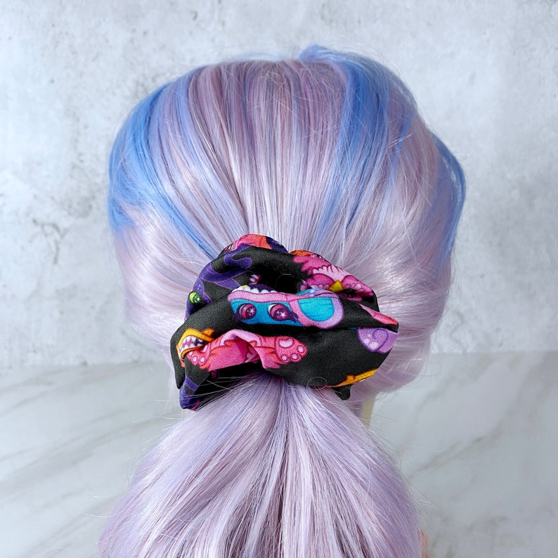 May include: A black hair scrunchie with a colorful print of cartoon monsters. The scrunchie is tied around a ponytail of purple and blue hair.