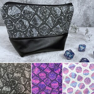 May include: A black zippered pouch with a white and silver design of dice. The pouch is made of a soft, textured fabric and has a black leather flap.