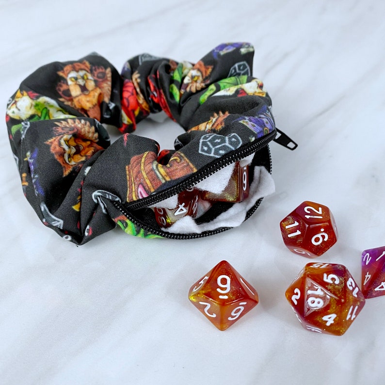 May include: A black fabric scrunchie with a zippered pocket and a colorful print of mythical creatures and dice. The scrunchie is open, revealing a white fleece lining and several red and orange dice with white numbers.