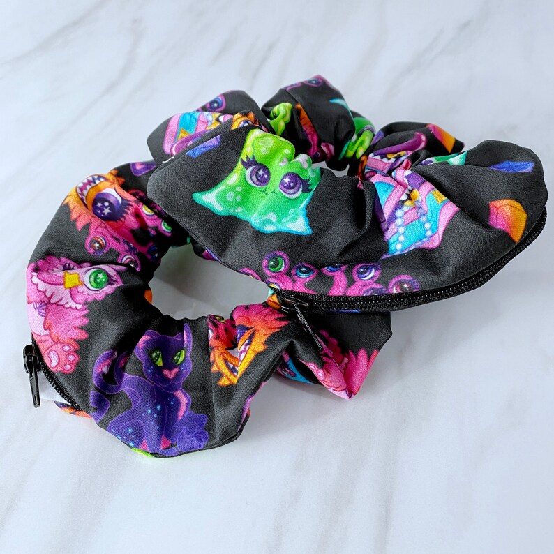 May include: Black scrunchie with a zipper closure and a colorful print of cartoon monsters and creatures.